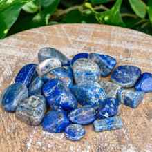 Load image into Gallery viewer, &quot;Pile of tumbled Lapis Lazuli crystals on a wooden plank with green foliage in the background,&quot; - Down to Earth.
