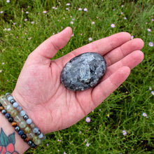Load image into Gallery viewer, &quot;Open hand with a large Larvikite palm stone in the center with greenery in the background,&quot; - Down to Earth.
