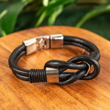 Load image into Gallery viewer, "Black braided leather bracelet with silver clasps on a wooden surface with green leaves in the background," - Down to Earth.

