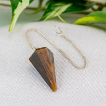 Load image into Gallery viewer, Tiger’s Eye Faceted Pendulum on a light shaded cloth with greenery in the background - Down to Earth
