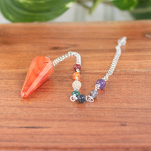 Load image into Gallery viewer, Premiere Carnelian Faceted Pendulum with 7 Chakra up close with plants on a style background - Down To Earth
