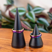 Load image into Gallery viewer, "Two black cone-shaped objects with colorful Rainbow Hematite bands on a wooden surface with a plant in the background," - Down to Earth.
