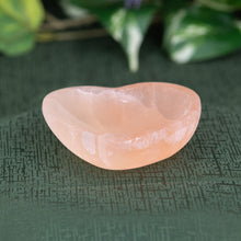 Load image into Gallery viewer, Red Selenite Heart-Shaped Bowl on a green textured surface with blurred purple flowers in the background front - Down to Earth
