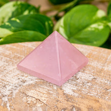 Load image into Gallery viewer, &quot;Pink Rose Quartz crystal on a wooden surface with green leaves in the background,&quot; - Down to Earth.
