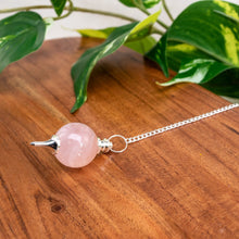 Load image into Gallery viewer, "Rose Quartz Sephoroton Pendulum on a wooden table with green foliage in the background," - Down to Earth.

