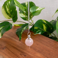 Load image into Gallery viewer, "Rose Quartz Sephoroton Pendulum hovering over a wooden table with green foliage in the background," - Down to Earth.
