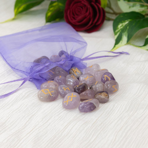Amethyst runes with a purple drawstring bag on a white surface with green leaves and a red rose in the background - Down to Earth
