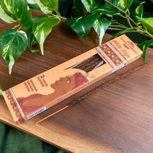 Load image into Gallery viewer, "Sacred Elements White Sage 10pk with a sole incense stick outside of the packaging on top of a wooden surface with green foliage in the background," - Down to Earth.
