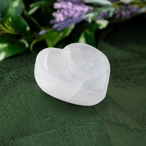 Selenite Heart-shaped Bowl on a green fabric background with blurred purple flowers in the background - Down to Earth