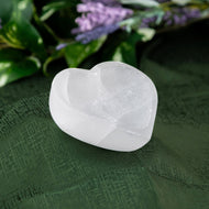 Selenite Heart-shaped Bowl on a green fabric background with blurred purple flowers in the background - Down to Earth