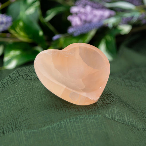 Red Selenite Heart-Shaped Bowl on a green textured surface with blurred purple flowers in the background - Down to Earth