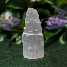 Load image into Gallery viewer, Selenite Tower on a textured green surface with blurred purple flowers in the background - Down to Earth
