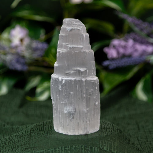 Selenite Tower on a textured green surface with blurred purple flowers in the background - Down to Earth