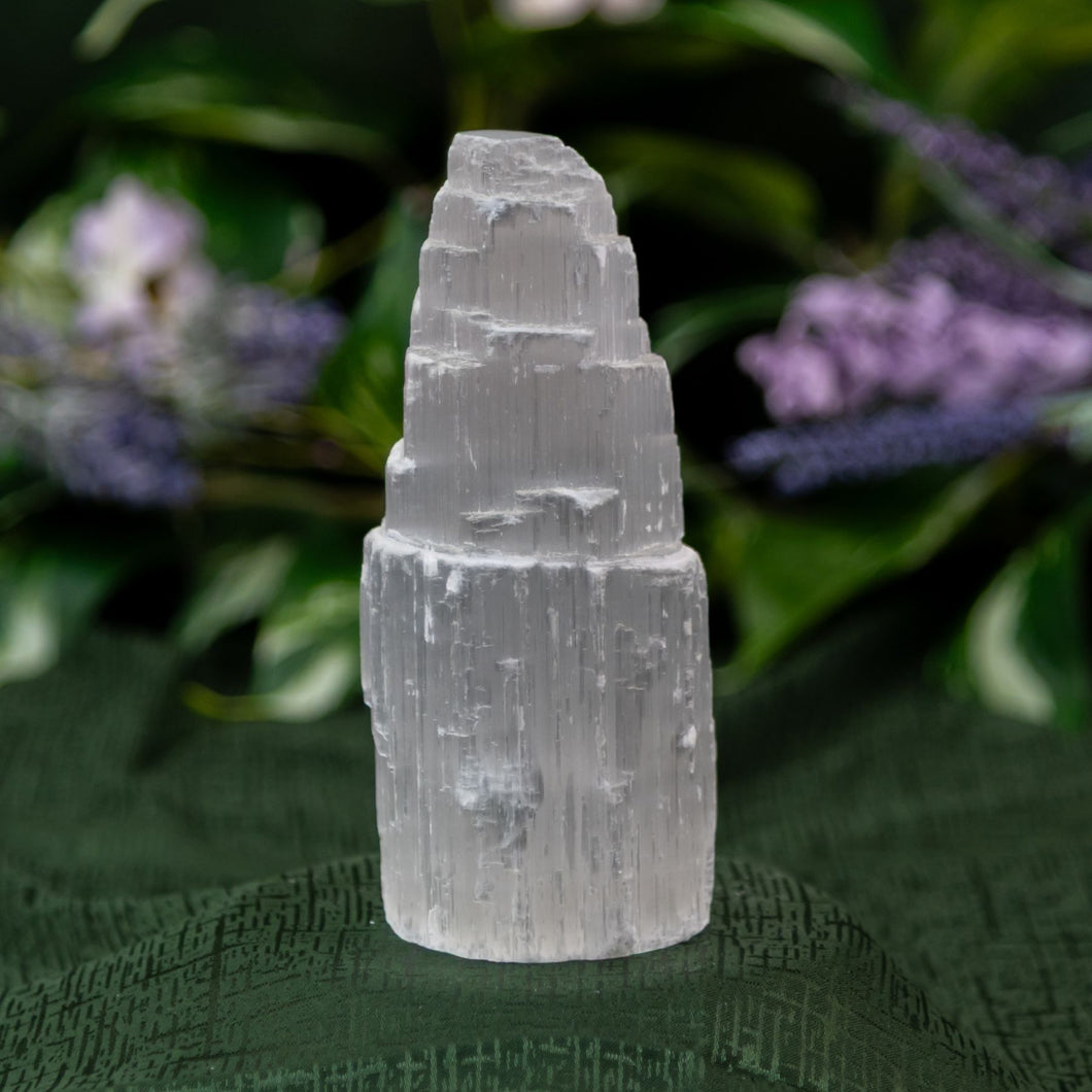 Selenite Tower on a textured green surface with blurred purple flowers in the background - Down to Earth