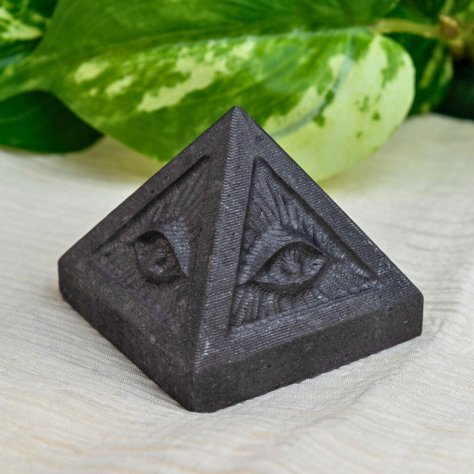 Black shungite pyramid-shaped object with evil eye design on a light surface with green leaves in the background - Down To Earth