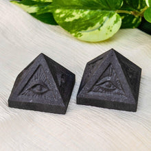 Load image into Gallery viewer, Two black shungite pyramid-shaped objects with evil eye designs on a light surface with green leaves in the background - Down To Earth
