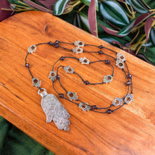 Load image into Gallery viewer, "Necklace with Hamsa hand pendant on a wooden surface with green leaves in the background," - Down to Earth.
