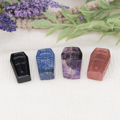 Four colorful crystal stones, including Black Obsidian, Sodalite, Amethyst, and Strawberry Quartz, on a white surface with blurred greenery in the background - Down to Earth