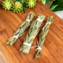 Load image into Gallery viewer, "Three bundles of Braided Sweet Grass on a wooden board with green foliage in the background," - Down to Earth.
