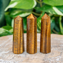 Load image into Gallery viewer, "Three Tiger's Eye Towers on a wooden base with green foliage in the background. This crystal is widely used in the metaphysical community to promote luck and abundance," - Down to Earth.

