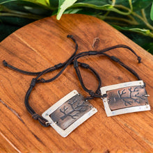 Load image into Gallery viewer, "Two metallic tree-stamped pendants on black cords against a wooden background with green leaves," - Down to Earth.
