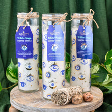 Load image into Gallery viewer, &quot;Three White Sage Evil Eye Pillar Candles on a small wooden table with green foliage in the background,&quot; - Down to Earth.
