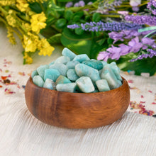 Load image into Gallery viewer, &quot;Wooden bowl overflowing with tumbled amazonite crystals on a cream-colored cloth against a floral background,&quot; - Down to Earth.
