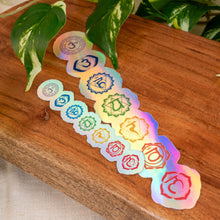 Load image into Gallery viewer, "Holographic stickers with chakra symbols on a wooden surface with green foliage in the background," - Down to Earth.
