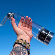 Load image into Gallery viewer, &quot;Clear water bottle with Black Obsidian chips in the bottom chamber held by a hand against a blue sky,&quot; - Down to Earth.
