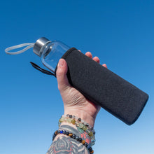 Load image into Gallery viewer, &quot;Hand holding a clear glass water bottle with a black sleeve against a blue sky,&quot; - Down to Earth.
