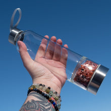 Load image into Gallery viewer, &quot;Clear water bottle with Carnelian crystal chips in the bottom chamber held in a hand against a clear blue sky,&quot; - Down to Earth.
