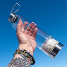 Load image into Gallery viewer, &quot;Clear glass water bottle with Clear Quartz crystal chips in the separate bottom chamber, held in a hand against a clear blue sky,&quot; - Down to Earth.
