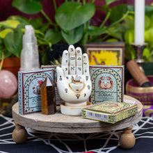 Load image into Gallery viewer, &quot;Decorative items including a palm reading backflow incense burner, zodiac incense cones, and crystals on a wooden stand with plants in the background,&quot; - Down to Earth.
