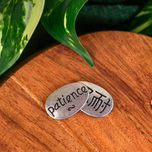 Load image into Gallery viewer, "Two silver oval stones with 'Patience' text on a wooden surface with green leaves in the background," - Down to Earth.

