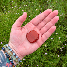 Load image into Gallery viewer, &quot;Open palm holding a 2&#39;&#39; tumbled Red jasper palm stone with green foliage in the background,&quot; - Down to Earth.
