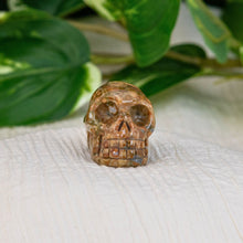 Load image into Gallery viewer, &quot;A carved rhyolite crystal skull on a cream-colored sheet with a greenery background,&quot; - Down to Earth.
