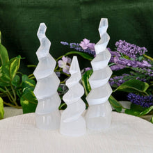Load image into Gallery viewer, &quot;Three selenite crystal towers on a white cloth with greenery and flowers in the background,&quot; - Down to Earth.

