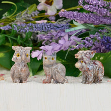 Load image into Gallery viewer, &quot;Three stone figurines on a white surface with purple flowers in the background,&quot; - Down to Earth.
