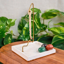 Load image into Gallery viewer, "Decorative rope incense burner with a twisted rope hanging from the golden metal hook on top of a wooden plank with green foliage in the background," - Down to Earth.

